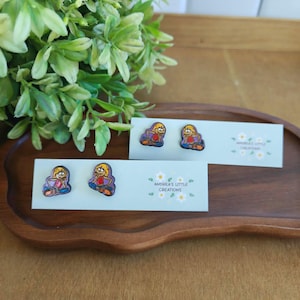 May include: Wooden stud earrings featuring a cartoon character with blonde hair, a red shirt, and a purple skirt. The earrings are displayed on a light blue card with the text "ANDREA'S LITTLE CREATIONS". The earrings are on a wooden tray.