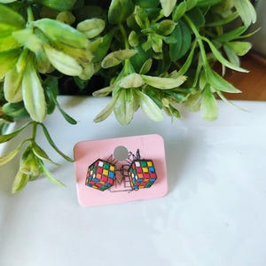 May include: A pair of colorful Rubik's Cube earrings on a pink card. The earrings feature a classic cube design with red, blue, yellow, green, orange, and white squares. The earrings are displayed on a white surface with green foliage in the background.