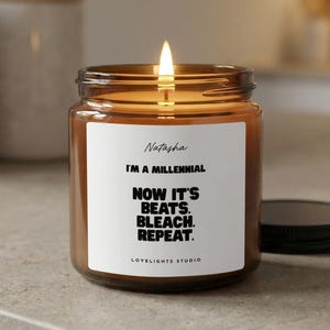 May include: A lit candle in a glass jar with a black lid. The label reads "Natasha, I'M A MILLENNIAL, NOW IT'S BEATS. BLEACH. REPEAT." The jar is brown and sits on a light-colored surface. The text "LOVELIGHTS STUDIO" is at the bottom of the label.