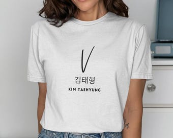 Op de afbeelding: Een wit t-shirt met korte mouwen en een zwarte grafische print op de borst. Het ontwerp bevat een grote letter V, de Koreaanse tekens 김태형 en de tekst KIM TAEHYUNG, gedrukt in een strak, gecentreerd lettertype zonder schreef.