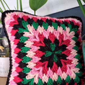 Pode incluir: Uma almofada de croch&ecirc; rosa, vermelha, verde e preta com um design quadrado. A almofada est&aacute; sentada em uma prateleira de madeira.