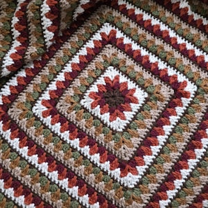 Puede incluir: Una manta de ganchillo hecha a mano con un patrón cuadrado. La manta presenta una paleta de colores marrón, blanco, verde y óxido. El diseño es una serie de cuadrados concéntricos, creando un textil texturizado y visualmente atractivo.