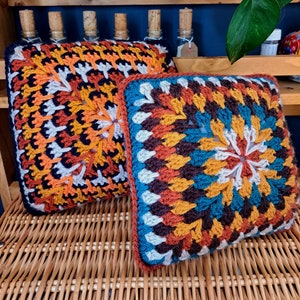 Op de afbeelding: Twee gehaakte kussens met een kleurrijk geometrisch patroon. De kussens zijn gemaakt van verschillende kleuren, waaronder oranje, bruin, blauw en wit. De kussens liggen op een rieten oppervlak.