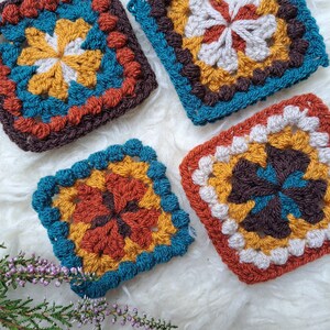 May include: Four crocheted squares in different color combinations. The squares feature a central flower design with a brown, white, yellow, and teal color scheme.