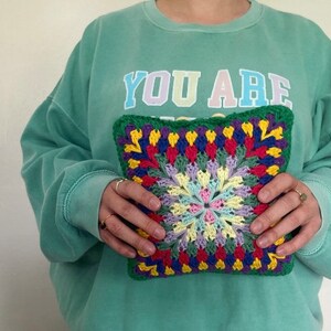 Pode incluir: Uma pessoa est&aacute; segurando um travesseiro quadrado de croch&ecirc; colorido com um fundo verde e um padr&atilde;o floral multicolorido no centro. O travesseiro est&aacute; sendo segurado na frente de um moletom verde com o texto "YOU ARE" impresso na frente.