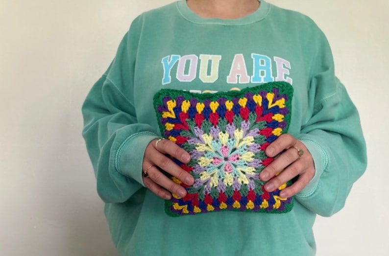 Pode incluir: Uma pessoa est&aacute; segurando um travesseiro quadrado de croch&ecirc; colorido com um fundo verde e um padr&atilde;o floral multicolorido no centro. O travesseiro est&aacute; sendo segurado na frente de um moletom verde com o texto "YOU ARE" impresso na frente.