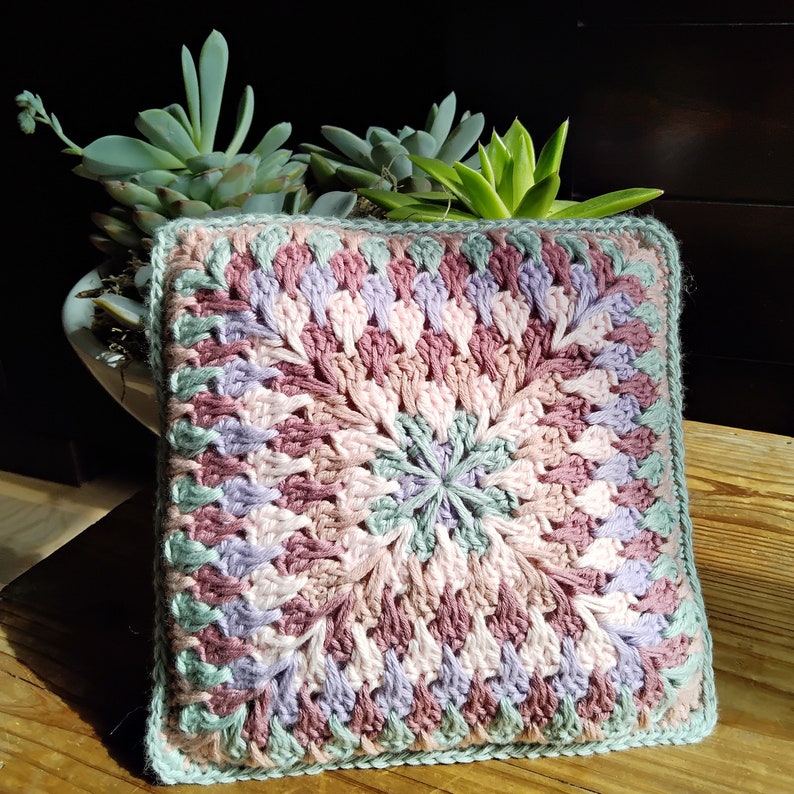 Pode incluir: Uma almofada quadrada de croch&ecirc; com um padr&atilde;o geom&eacute;trico multicolorido em tons de rosa, verde e roxo. A almofada &eacute; feita com um fio grosso e tem uma superf&iacute;cie texturizada.