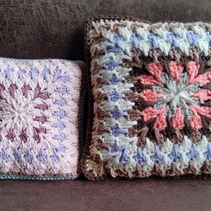 Pode incluir: Duas almofadas quadradas de croch&ecirc; com padr&otilde;es de cores diferentes. A almofada maior &eacute; marrom, branca e azul claro com um centro rosa. A almofada menor &eacute; rosa, roxa e branca com uma borda azul claro.