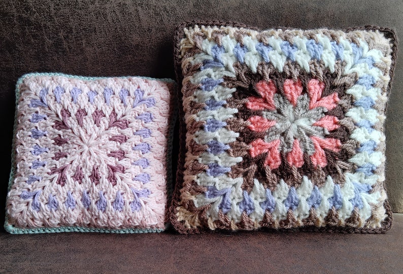 Pode incluir: Duas almofadas quadradas de croch&ecirc; com padr&otilde;es de cores diferentes. A almofada maior &eacute; marrom, branca e azul claro com um centro rosa. A almofada menor &eacute; rosa, roxa e branca com uma borda azul claro.
