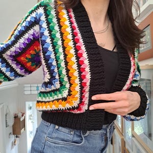 May include: A black crochet cardigan with a rainbow colored trim. The cardigan is cropped and has a V-neck.