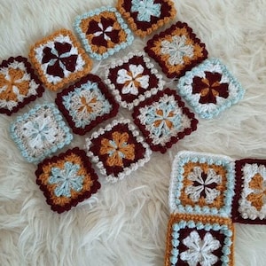 May include: A collection of crocheted squares in various colors, including white, brown, blue, and yellow. The squares are arranged in a pattern and feature a flower design in the center.