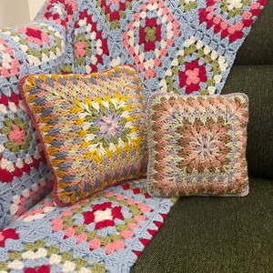 Pode incluir: Dois almofadas de sof&aacute; de croch&ecirc; com um padr&atilde;o de quadrados da av&oacute; em tons de rosa, azul, verde e amarelo. As almofadas est&atilde;o sentadas em um sof&aacute; coberto com uma manta de croch&ecirc; de quadrados da av&oacute; azuis e verdes.