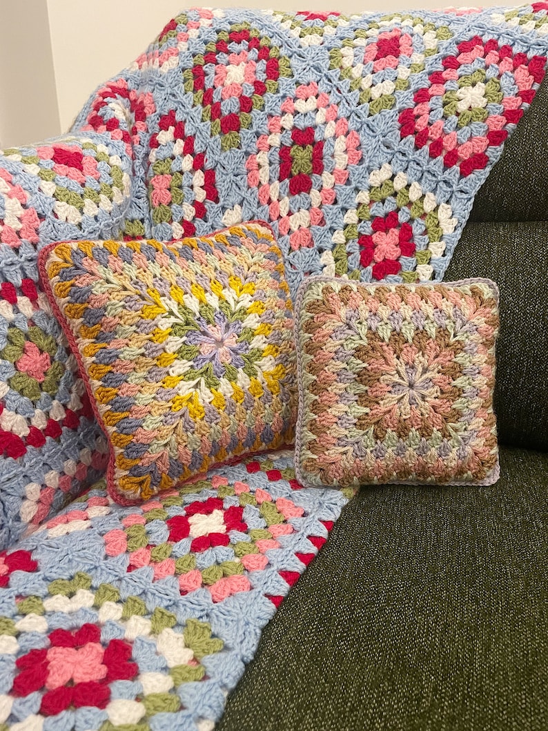 Pode incluir: Dois almofadas de sof&aacute; de croch&ecirc; com um padr&atilde;o de quadrados da av&oacute; em tons de rosa, azul, verde e amarelo. As almofadas est&atilde;o sentadas em um sof&aacute; coberto com uma manta de croch&ecirc; de quadrados da av&oacute; azuis e verdes.