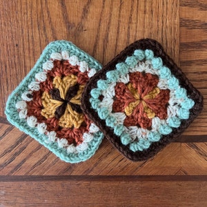 May include: Two crocheted squares with a brown, white, and orange floral pattern. The squares are made with a light blue yarn and have a brown border.