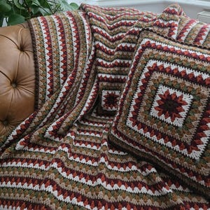 May include: A crocheted blanket and pillow set in a geometric pattern. The blanket and pillow feature a repeating chevron design in shades of brown, red, green, and white. The pillow is square with a central floral motif.