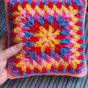 Pode incluir: Uma almofada quadrada de croch&ecirc; com um padr&atilde;o geom&eacute;trico colorido em tons de rosa, vermelho, amarelo e azul.