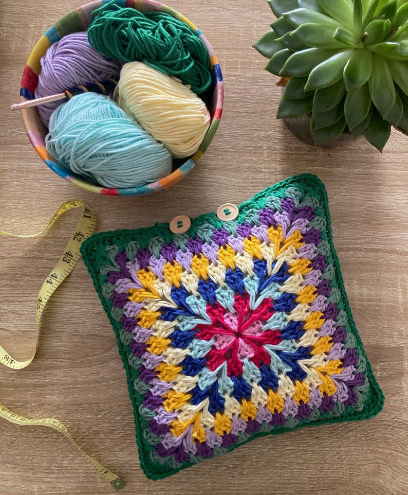 Pode incluir: Uma almofada de croch&ecirc; colorida com uma borda verde e um padr&atilde;o geom&eacute;trico multicolorido no centro. A almofada &eacute; feita com diferentes tons de fio roxo, azul, amarelo e rosa.