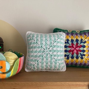 Pode incluir: Duas almofadas quadradas de croch&ecirc;, uma em verde e branco com um padr&atilde;o em espiral, e a outra em um padr&atilde;o floral multicolorido com uma borda verde.