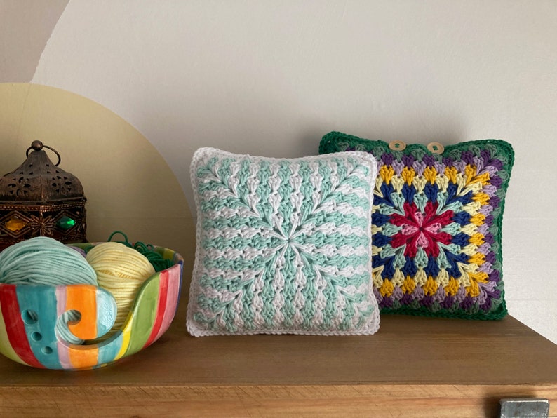 Pode incluir: Duas almofadas quadradas de croch&ecirc;, uma em verde e branco com um padr&atilde;o em espiral, e a outra em um padr&atilde;o floral multicolorido com uma borda verde.