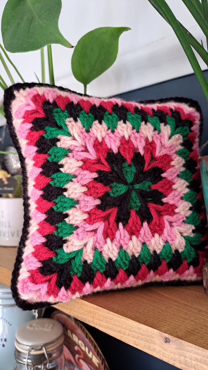Pode incluir: Uma almofada de croch&ecirc; rosa, vermelha, verde e preta com um design quadrado. A almofada est&aacute; sentada em uma prateleira de madeira.