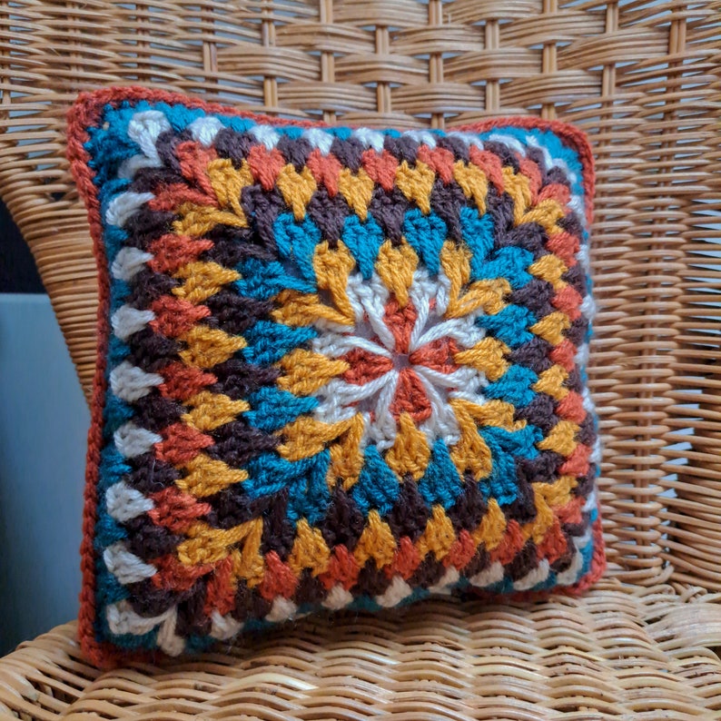 Pode incluir: Uma almofada de croch&ecirc; colorida com um padr&atilde;o geom&eacute;trico em tons de laranja, marrom, azul-turquesa e branco. A almofada est&aacute; sentada em uma cadeira de vime.