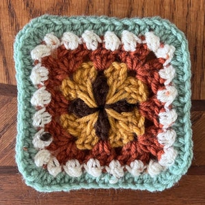 May include: A crocheted square with a flower design in shades of brown, orange, and white. The square is surrounded by a border of green and white.