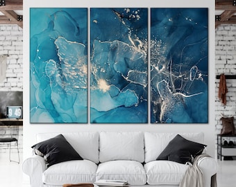 May include: A triptych of abstract art panels featuring flowing blue and gold designs. The artwork has a fluid, watercolor-like appearance with varying shades of blue and shimmering gold accents. The panels are displayed above a white sofa with black pillows.