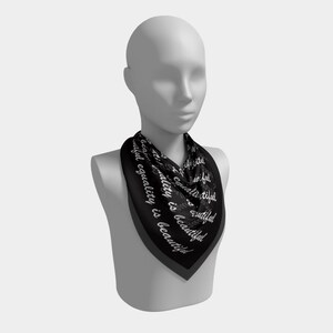 May include: A black square scarf with the words "beautiful equality is beautiful" in white script. The scarf is draped around a mannequin's neck, showcasing its design and shape. The scarf has a dark gray border.