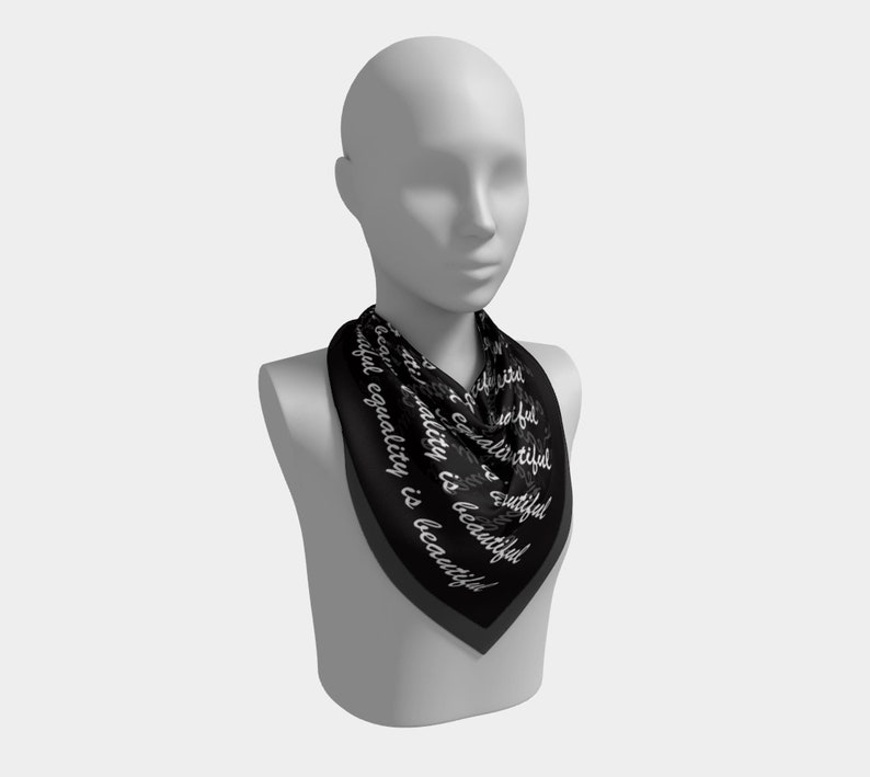 May include: A black square scarf with the words "beautiful equality is beautiful" in white script. The scarf is draped around a mannequin's neck, showcasing its design and shape. The scarf has a dark gray border.