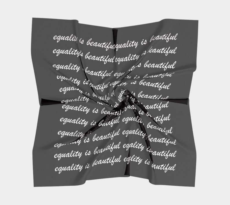 May include: A dark gray square scarf with the words "equality is beautiful" repeated in white script across the fabric. The scarf is twisted and folded, creating a dynamic visual effect.