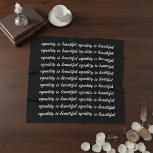 May include: A black textile with the phrase "equality is beautiful" repeated in white cursive script. The textile is displayed on a dark wooden surface, accompanied by a stack of books, a teacup, and decorative items.