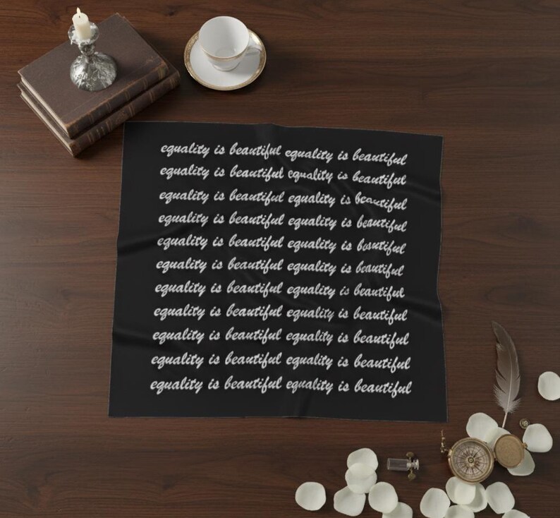 May include: A black textile with the phrase "equality is beautiful" repeated in white cursive script. The textile is displayed on a dark wooden surface, accompanied by a stack of books, a teacup, and decorative items.