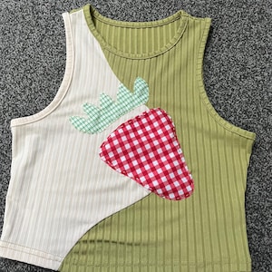 Pode incluir: Um top curto sem mangas com um design único. O top apresenta um painel creme à esquerda, um painel verde à direita e uma aplicação de morango. O morango é xadrez vermelho e branco com folhas xadrez verdes.
