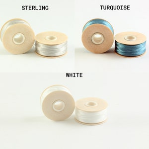 May include: Three spools of thread in different colors: sterling, turquoise, and white. Each spool is made of wood and has a hole in the center for threading.