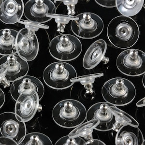 May include: A close-up of a group of clear plastic earring backs. The backs have a small metal post that goes through the earring and a flat, round back that sits against the earlobe.