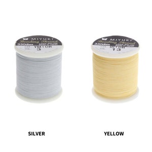 Op de afbeelding: Twee spoelen Miyuki Beading Thread, een zilver en een geel. De spoelen zijn gelabeld met de merknaam, het type draad, de maat en de kleur.
