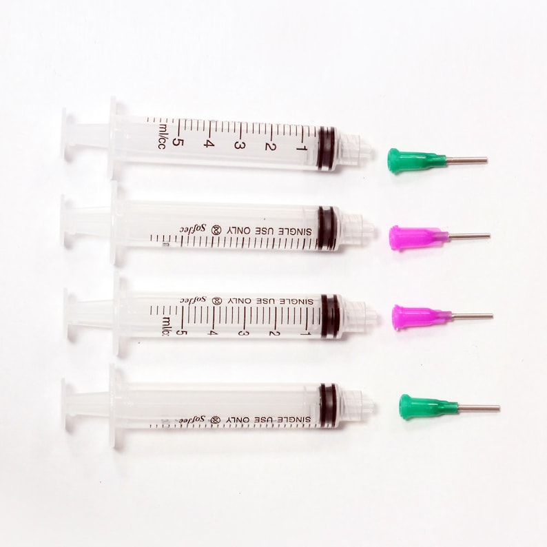 May include: Four clear plastic syringes with black rubber stoppers and measurement markings. Two syringes have green needle caps, and two have pink needle caps. The syringes are labelled "SINGLE USE ONLY" and "Softec".