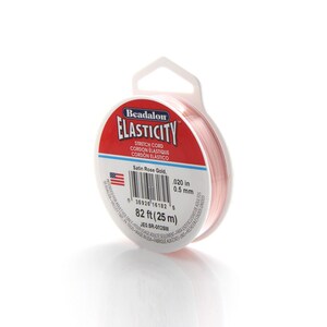 Może przedstawiać: Szpula r&oacute;żowego elastycznego sznurka do tworzenia biżuterii. Szpula jest oznaczona "Beadalon Elasticity Stretch Cord Cordon Elastique Cordon Elastico" i "Satin Rose Gold, 0.020 in 0.5 mm 82 ft (25 m)"