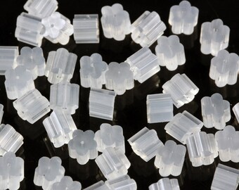 May include: A close-up of a pile of clear plastic earring backs. The backs are shaped like a flower with five petals and have a small hole in the centre. They are designed to be used with stud earrings.