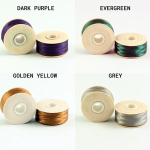May include: Four spools of thread in different colors: dark purple, evergreen, golden yellow, and grey. Each spool is made of wood and has a clear plastic center.
