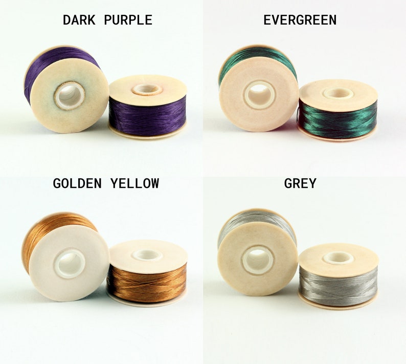 May include: Four spools of thread in different colors: dark purple, evergreen, golden yellow, and grey. Each spool is made of wood and has a clear plastic center.