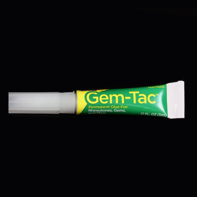 May include: A green and yellow tube of Gem-Tac permanent glue for rhinestones, gems, and more. The tube is labeled "Non-Toxic" and contains 0.17 fluid ounces (5 ml) of glue.