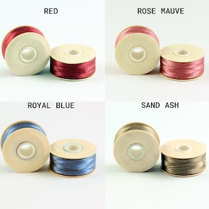 May include: Four spools of thread in different colors: red, rose mauve, royal blue, and sand ash. Each spool is wound with thread and has a white plastic bobbin.
