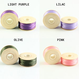 May include: Four spools of thread in different colors: light purple, lilac, olive, and pink. Each spool is made of wood and has a hole in the center for threading.