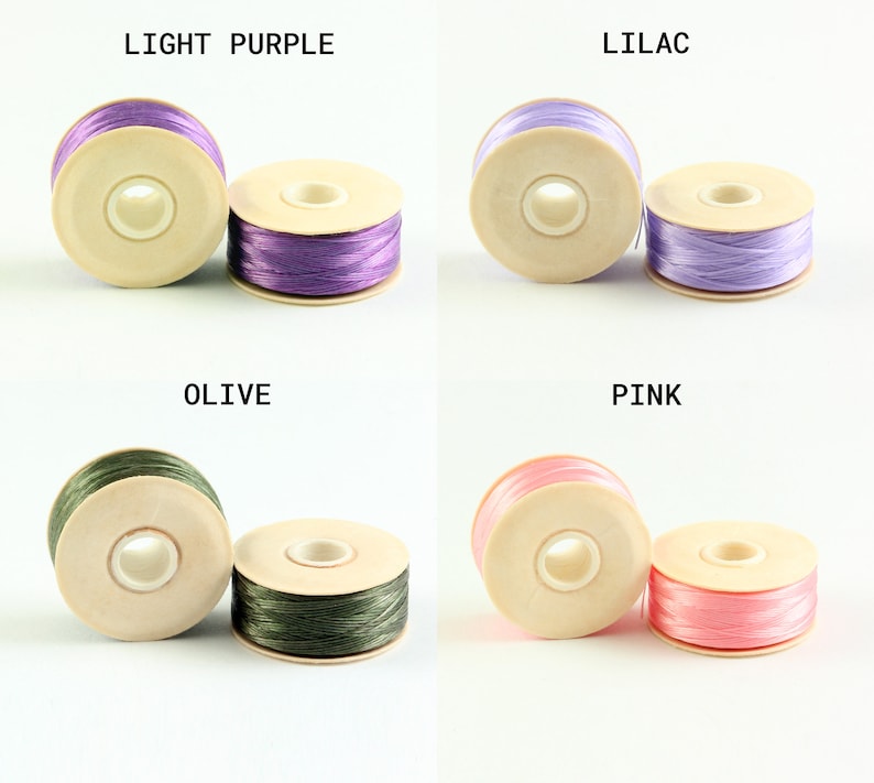 May include: Four spools of thread in different colors: light purple, lilac, olive, and pink. Each spool is made of wood and has a hole in the center for threading.