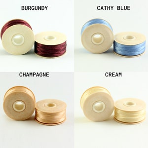 May include: Four spools of thread in different colors: burgundy, blue, champagne, and cream. Each spool is wound with thread and has a wooden base.