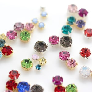 Op de afbeelding: Een verzameling kleurrijke strass-steentjes in verschillende maten en tinten, waaronder rood, roze, groen, blauw en bruin. Elke strass-steen heeft een zilveren of goudkleurige metalen zetting, waardoor een sprankelend effect ontstaat tegen een witte achtergrond.