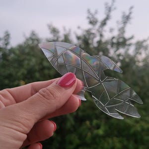 Op de afbeelding: Een doorschijnende, raketvormige sticker met een geometrisch, op glas-in-lood geïnspireerd patroon met iriserende, regenboogkleurige accenten die over het oppervlak verschuiven en een prismatisch effect creëren in het licht.