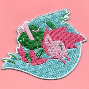 May include: Embroidered patch featuring a green frog and a pink hedgehog with closed eyes, surrounded by pink and green leaves on a light blue background.