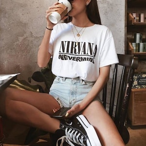 May include: A woman wearing a white t-shirt with the words "NIRVANA NEVERMIND" printed on it, blue denim shorts, and black high-top sneakers. She is holding a cup of coffee and sitting on a chair.